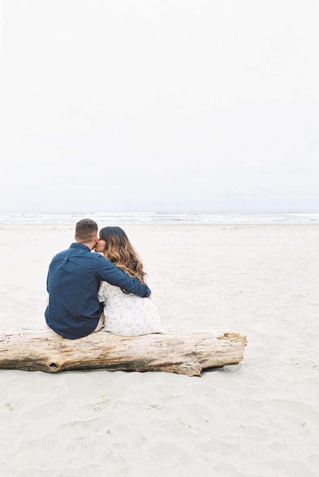Couple sitting on driftwood log, embracing and kissing while facing ocean on sandy beach