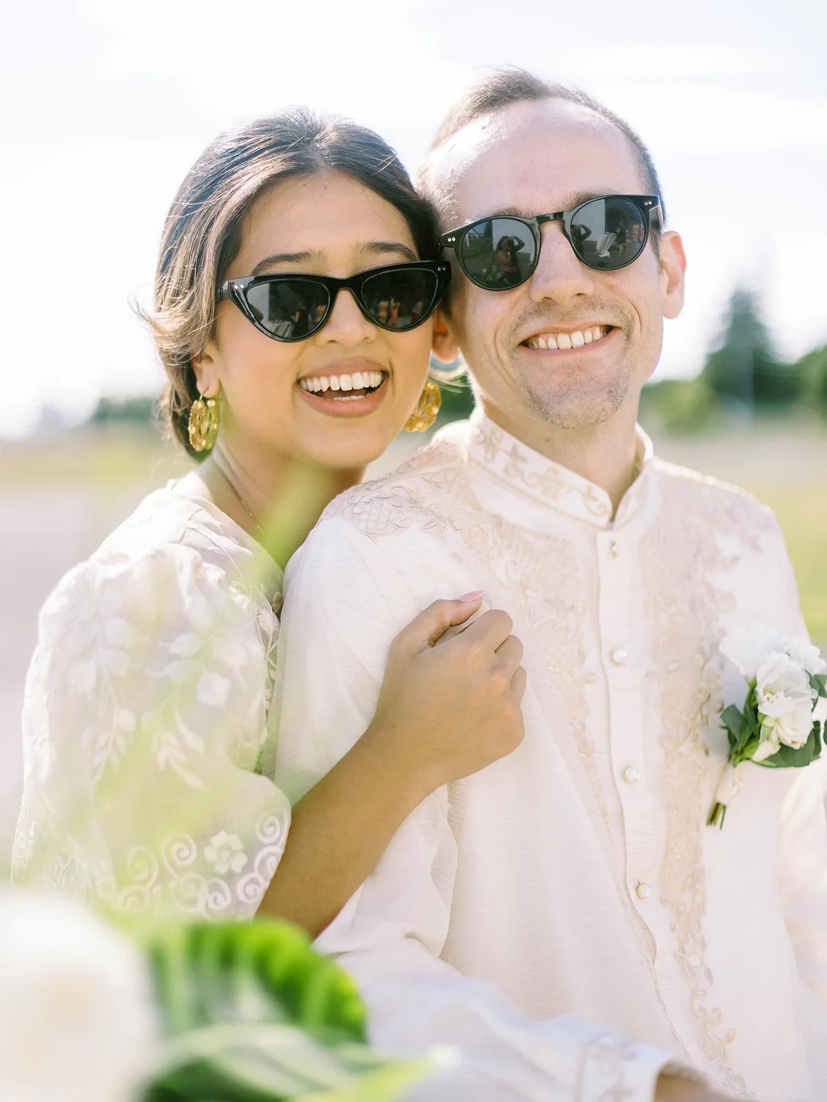 Close-up portrait of bride and groom both wearing sunglasses and smiling broadly at the camera in a sunny field
