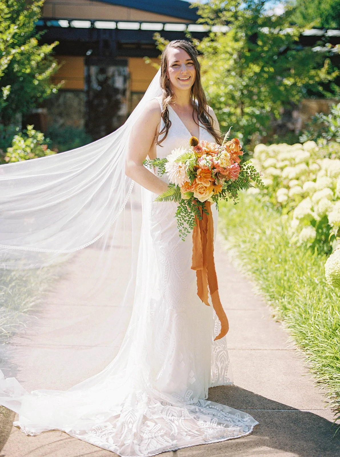 Bride standing on garden path with bouquet and long veil trailing behind