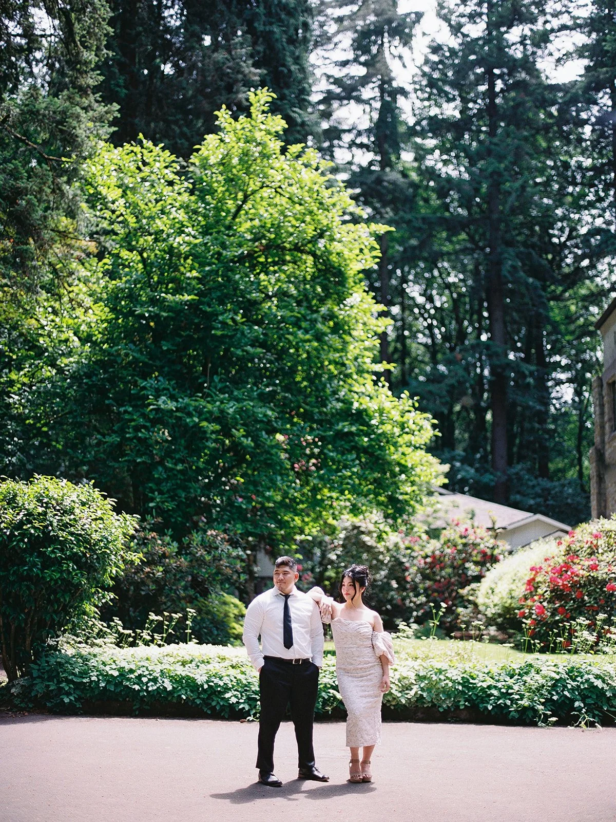 Wide shot of the bride and groom standing side by side on a sun-drenched path at The Grotto, towering evergreens and blooming rhododendrons behind them