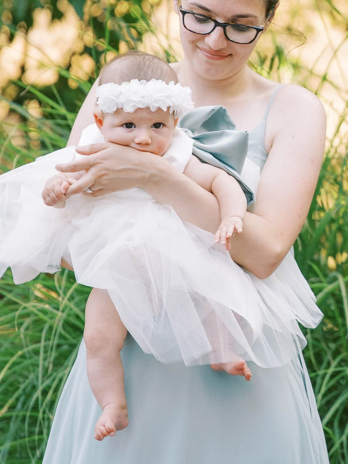 Baby flower girl in white dress with floral headband held by bridesmaid at outdoor wedding.