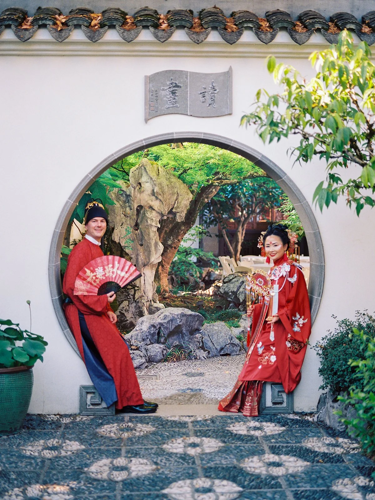 Bride and groom posing together inside a circular moon gate, both smiling at the camera with a pebbled garden courtyard in the foreground
