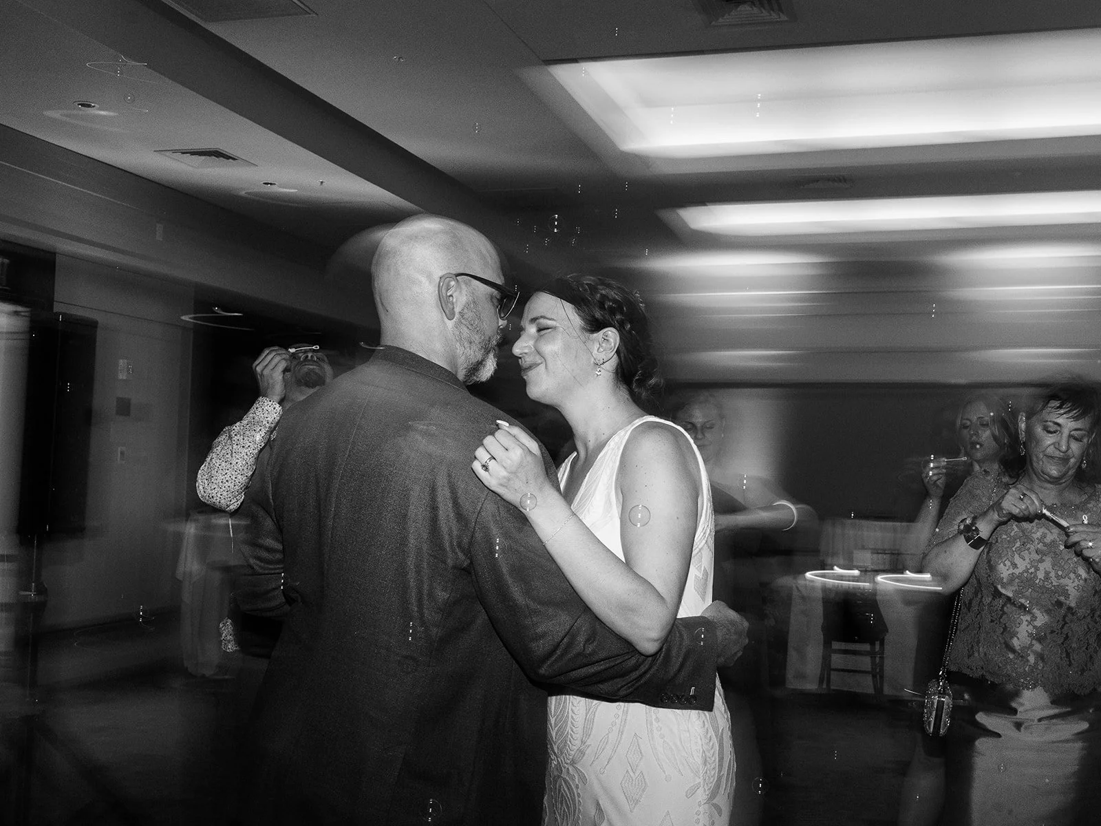 Black and white photo of the bride smiling up at the groom as they slow dance, surrounded by floating bubbles and guests