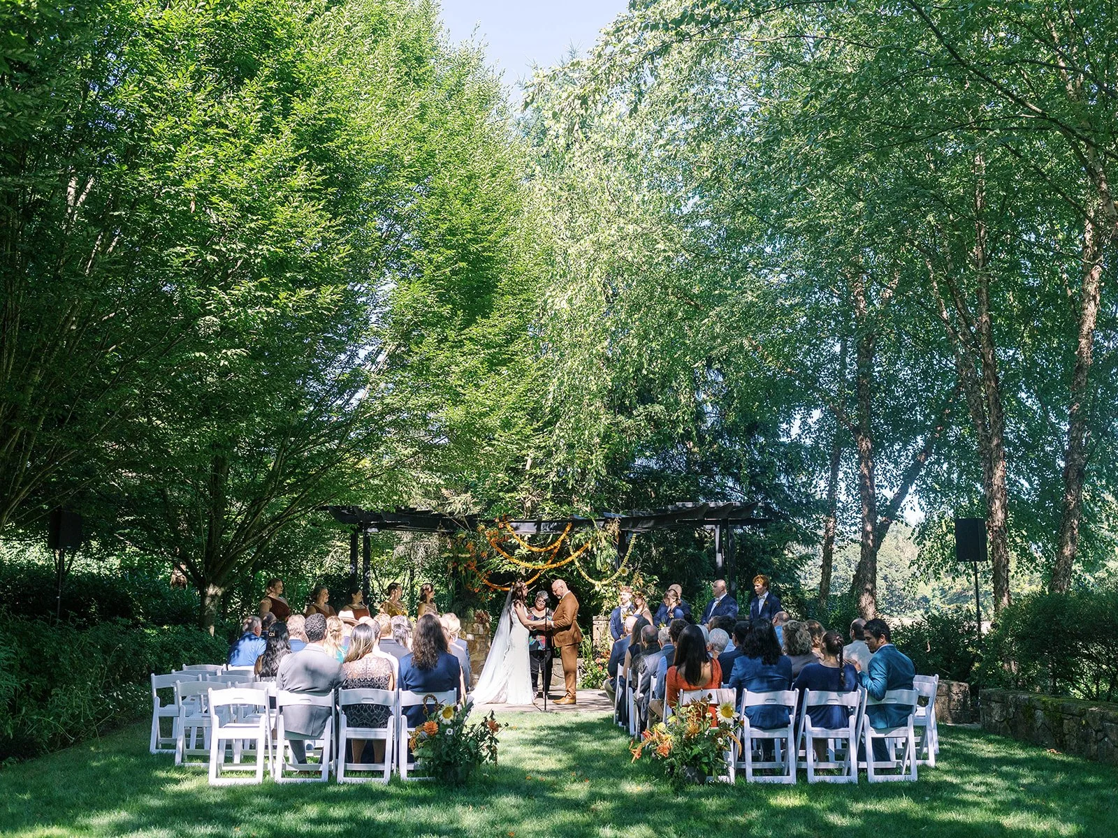 Wide view of outdoor wedding ceremony with guests seated on white chairs facing a floral-decorated arbor.