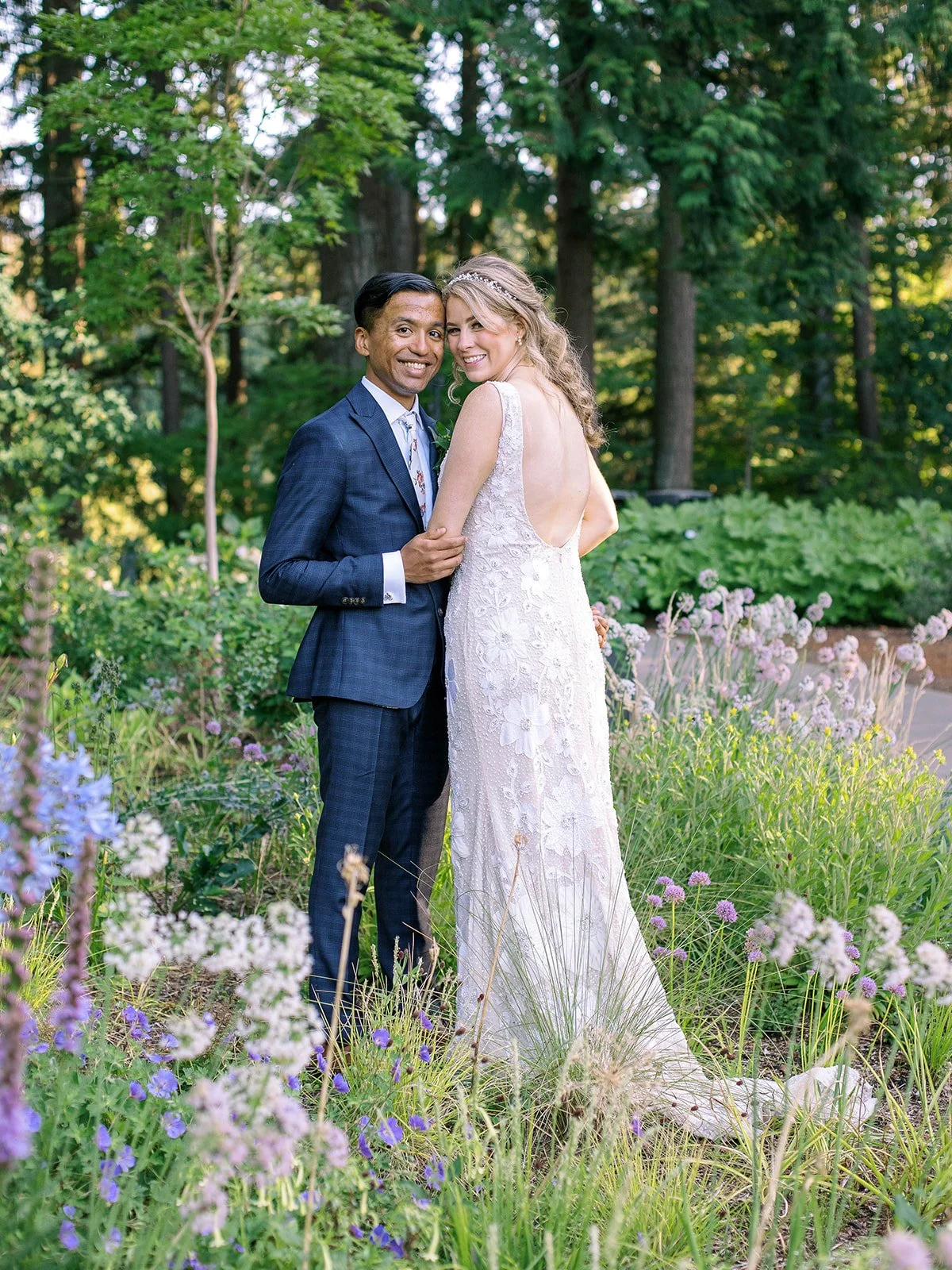 Bride and groom pose among purple wildflowers and lush garden greenery.