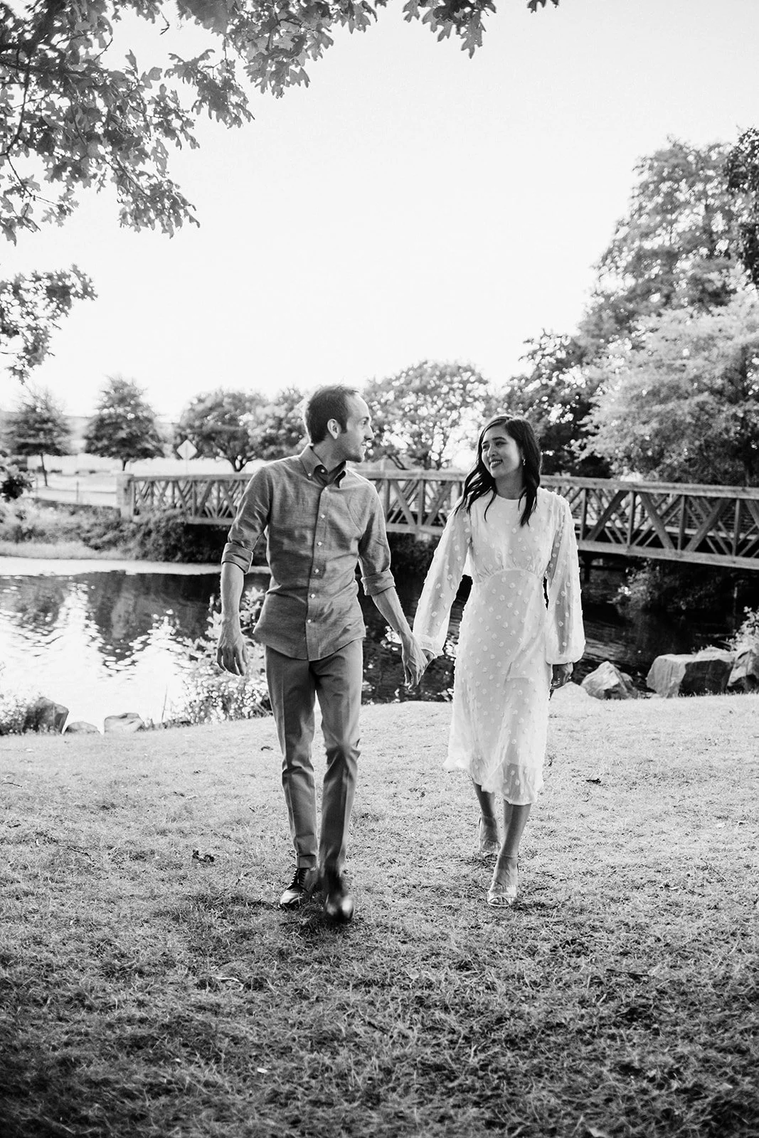 Black and white image of couple holding hands walking near a wooden bridge over water in a park setting