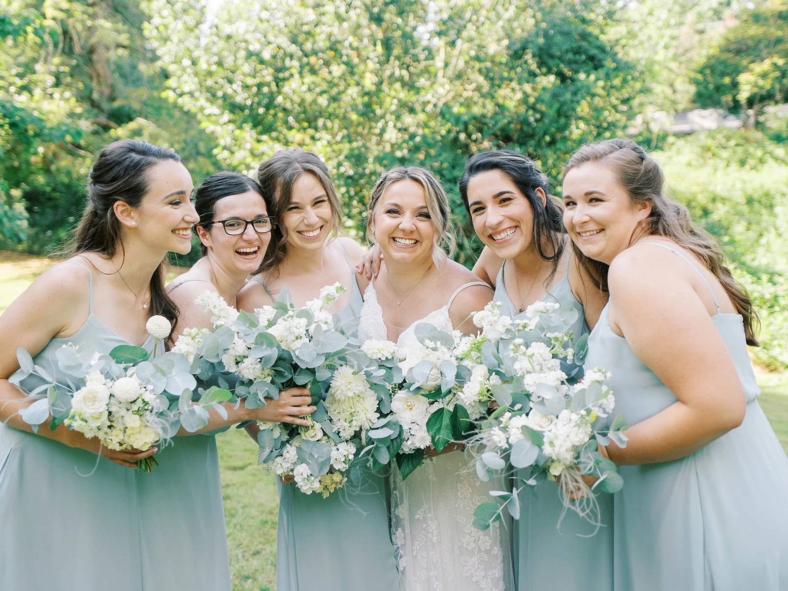 Bride smiling with bridesmaids in sage dresses holding lush white floral bouquets outdoors.