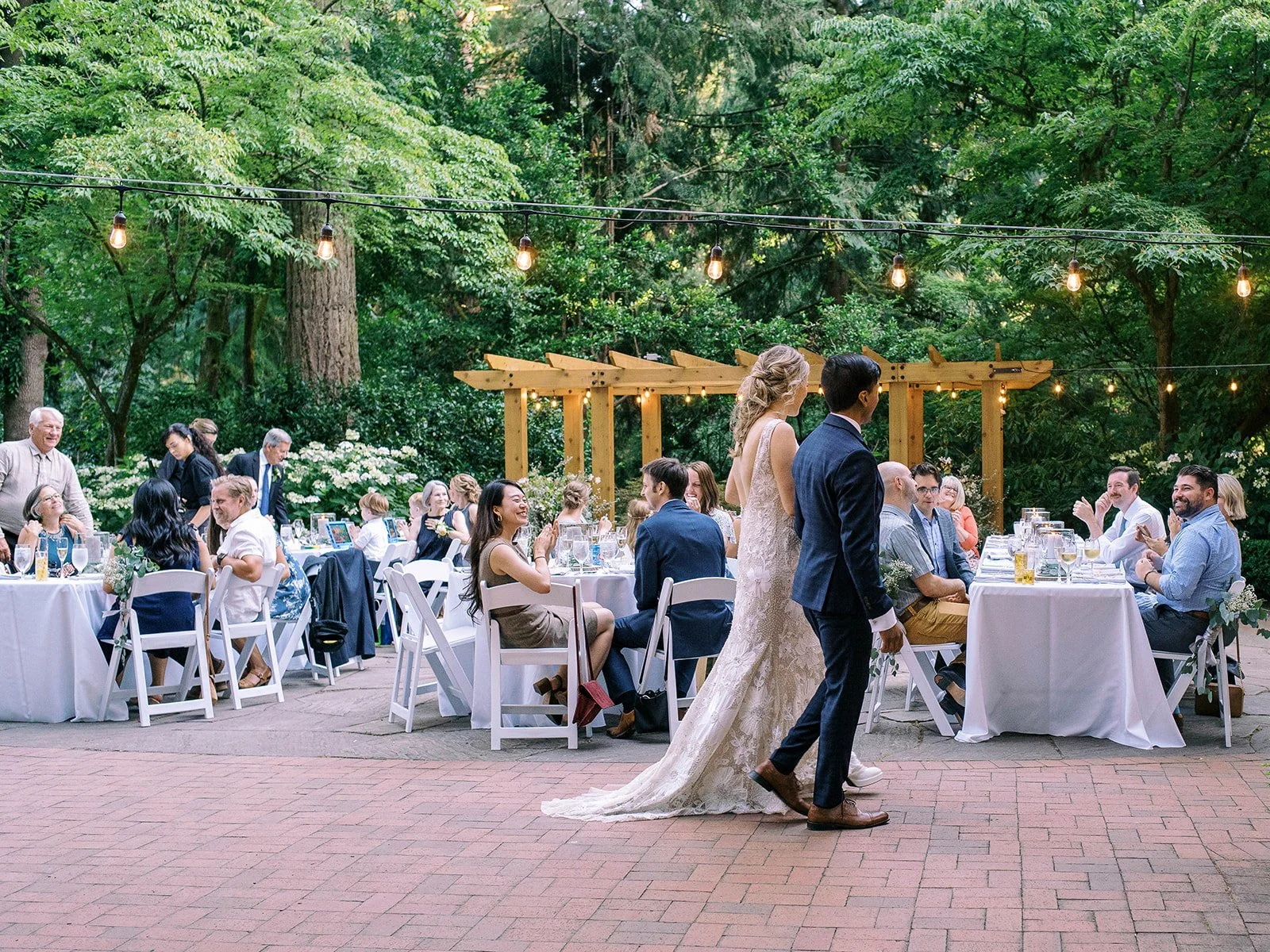 Newlyweds walk through their outdoor reception beneath string lights and surrounding trees.