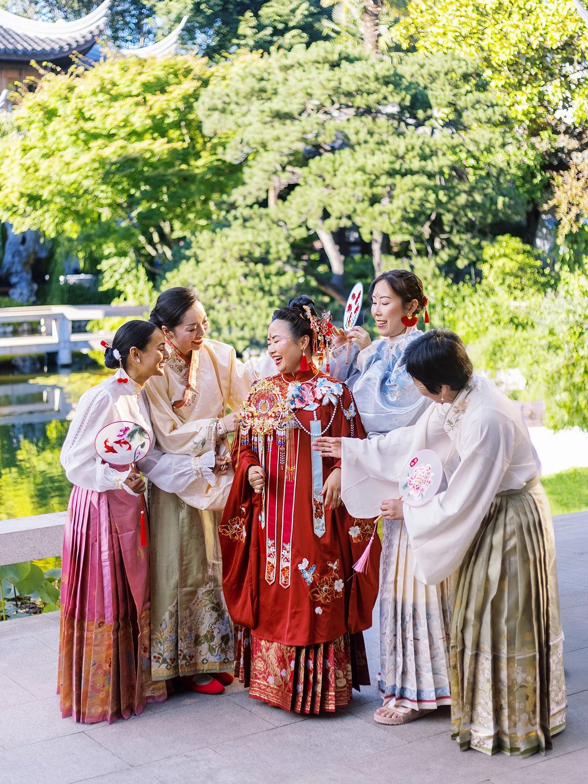 Bride in red traditional attire surrounded by attendants in pastel hanfu adjusting her accessories in a garden setting