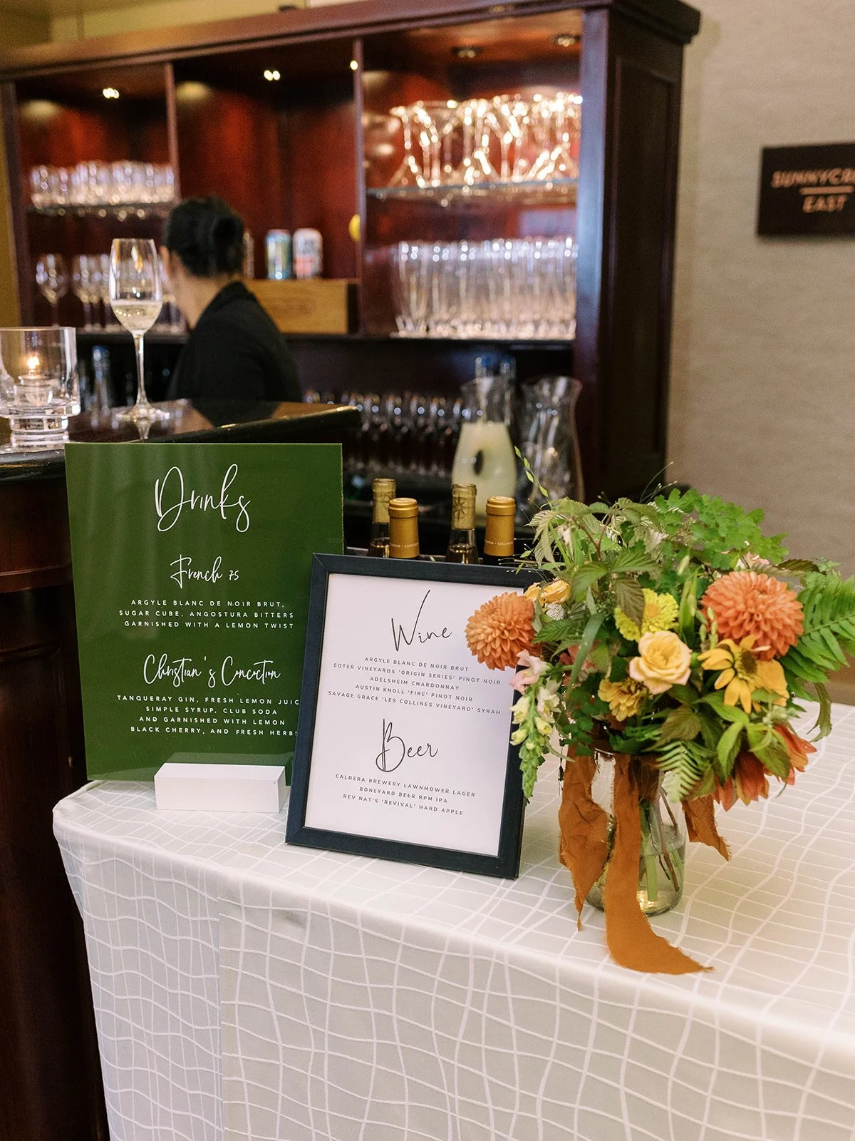 Wedding welcome display with sign, floral arrangements, and card box on a wooden table in reception space.