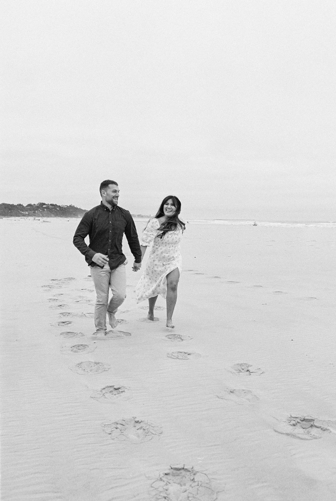 Couple walking hand in hand along beach with footprints trailing behind, black and white photo