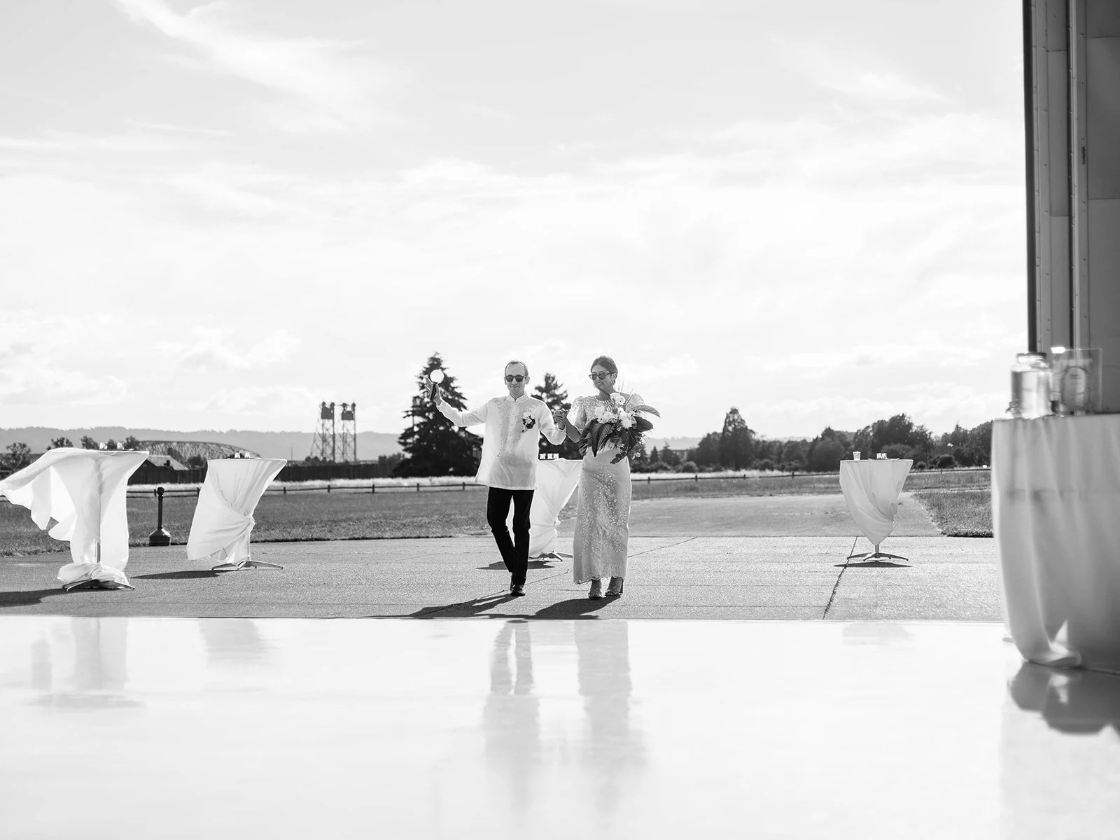 Black and white wide shot of bride and groom entering the Pearson Air Museum hangar in sunglasses, groom raising a drink in celebration
