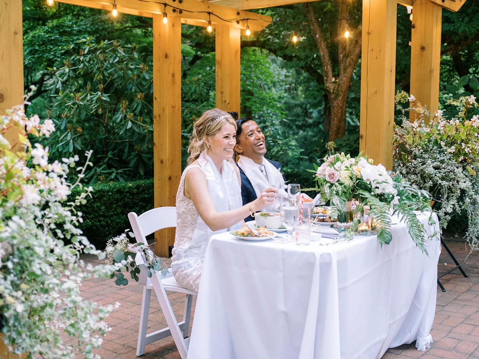 Bride and groom laugh together at their flower-filled sweetheart table.