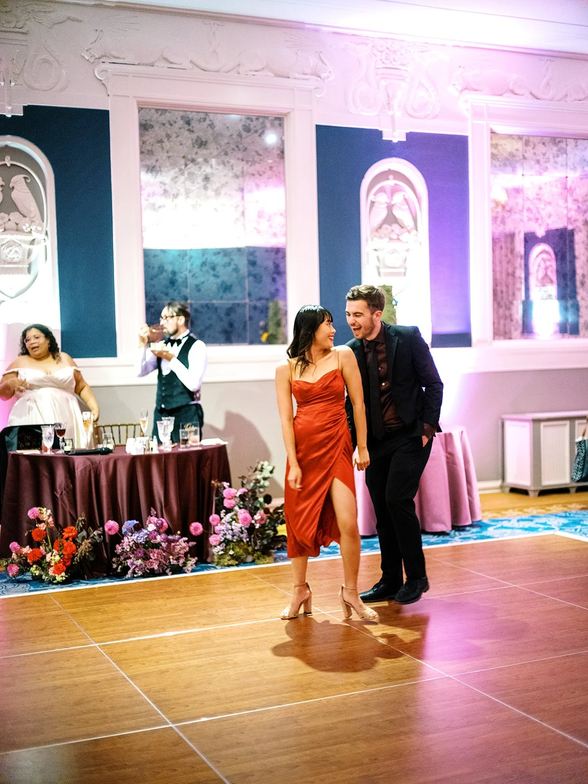 Couple dancing together on wooden dance floor with guests and floral sweetheart table in background.