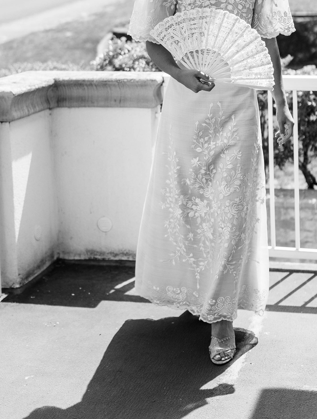 Black and white image of the bride's embroidered gown skirt and heeled sandals while holding a lace fan on a terrace
