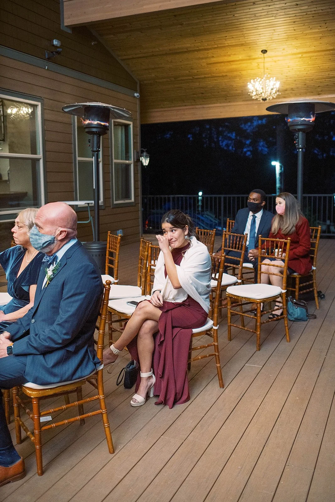 Wedding guest wipes her eye while seated beneath patio heaters during the intimate evening ceremony.