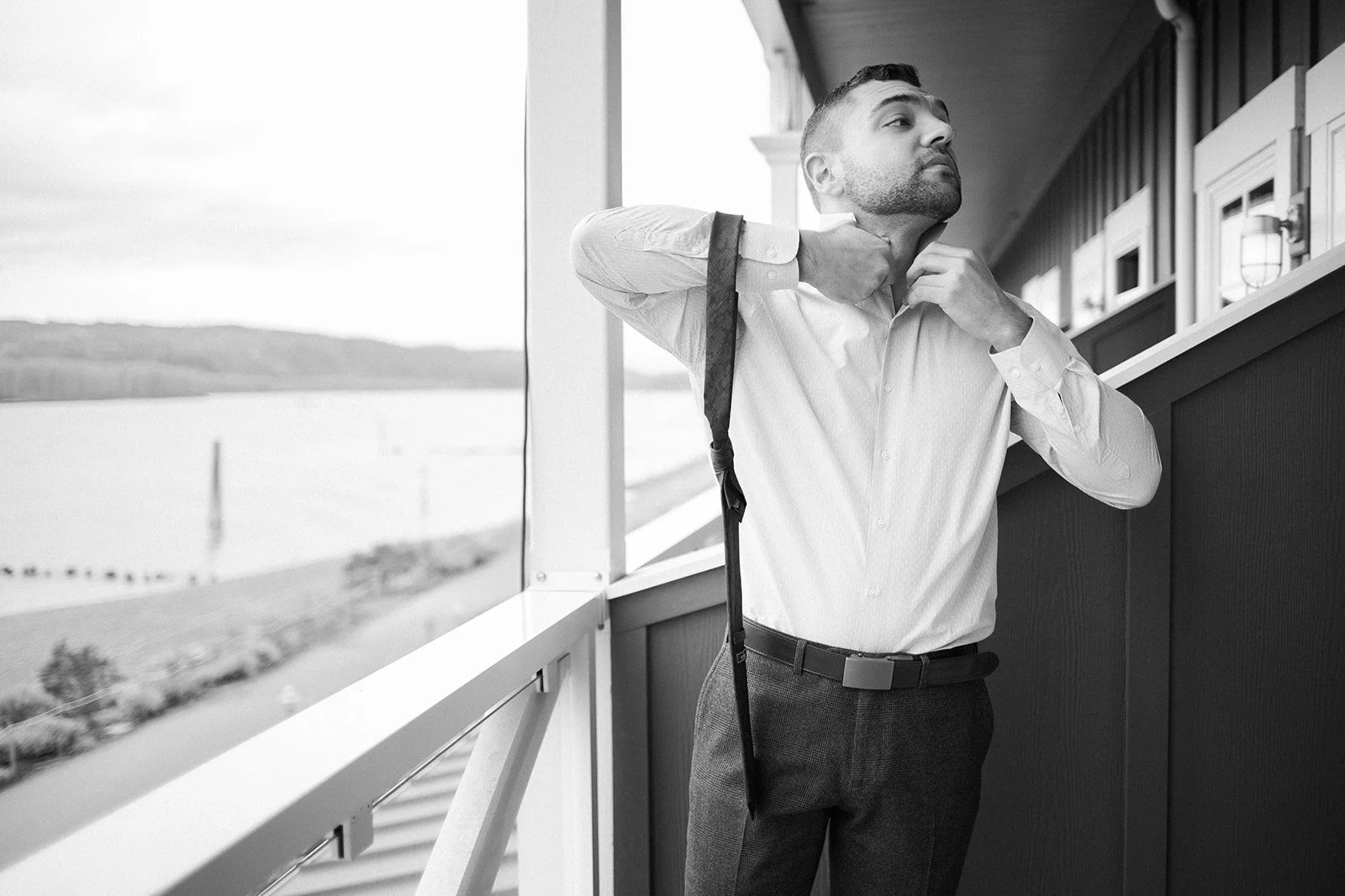Black-and-white portrait of the groom adjusting his shirt while getting ready beside the river.