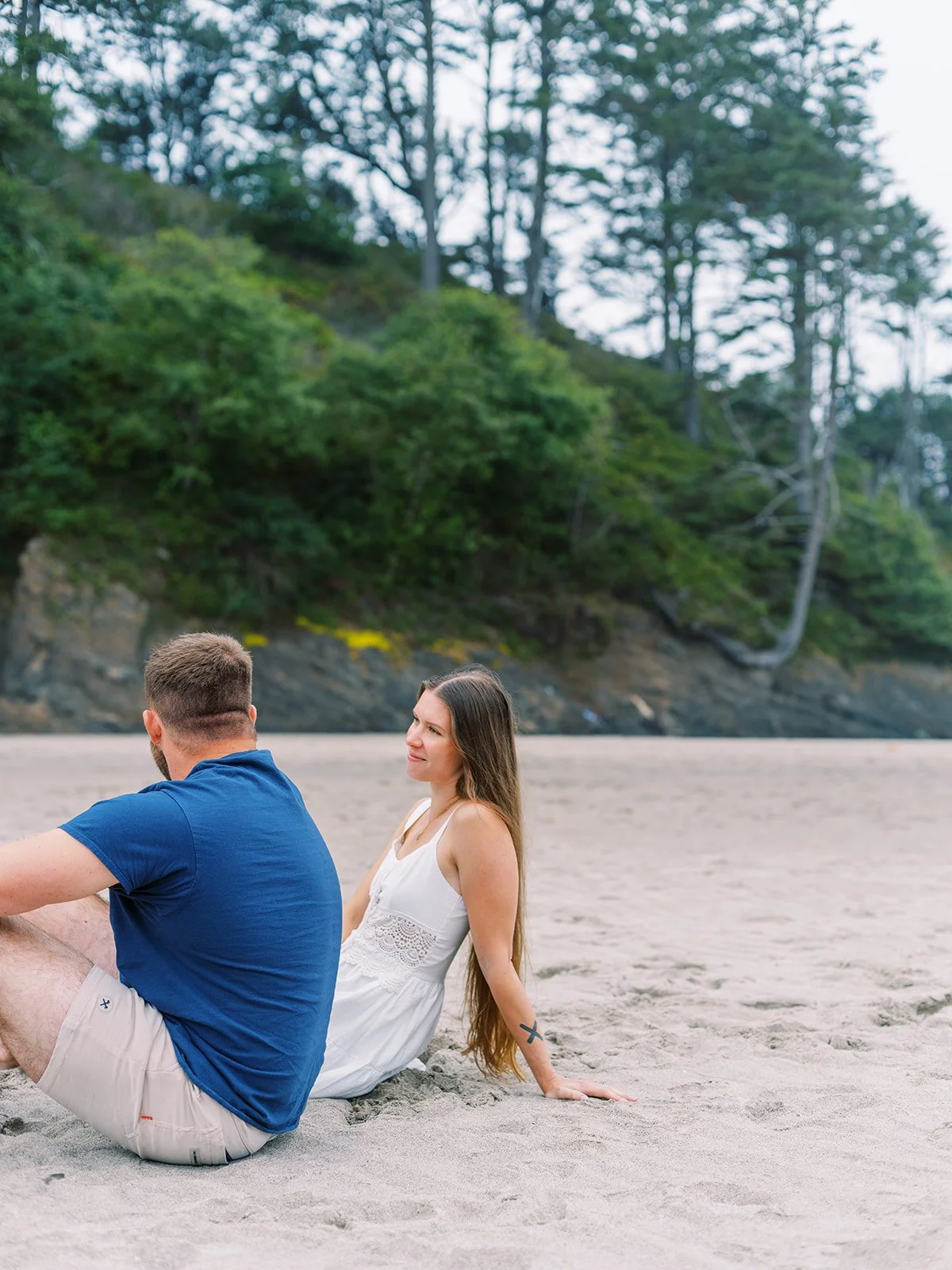 Couple sitting on a sandy beach with green hillside and trees behind them.