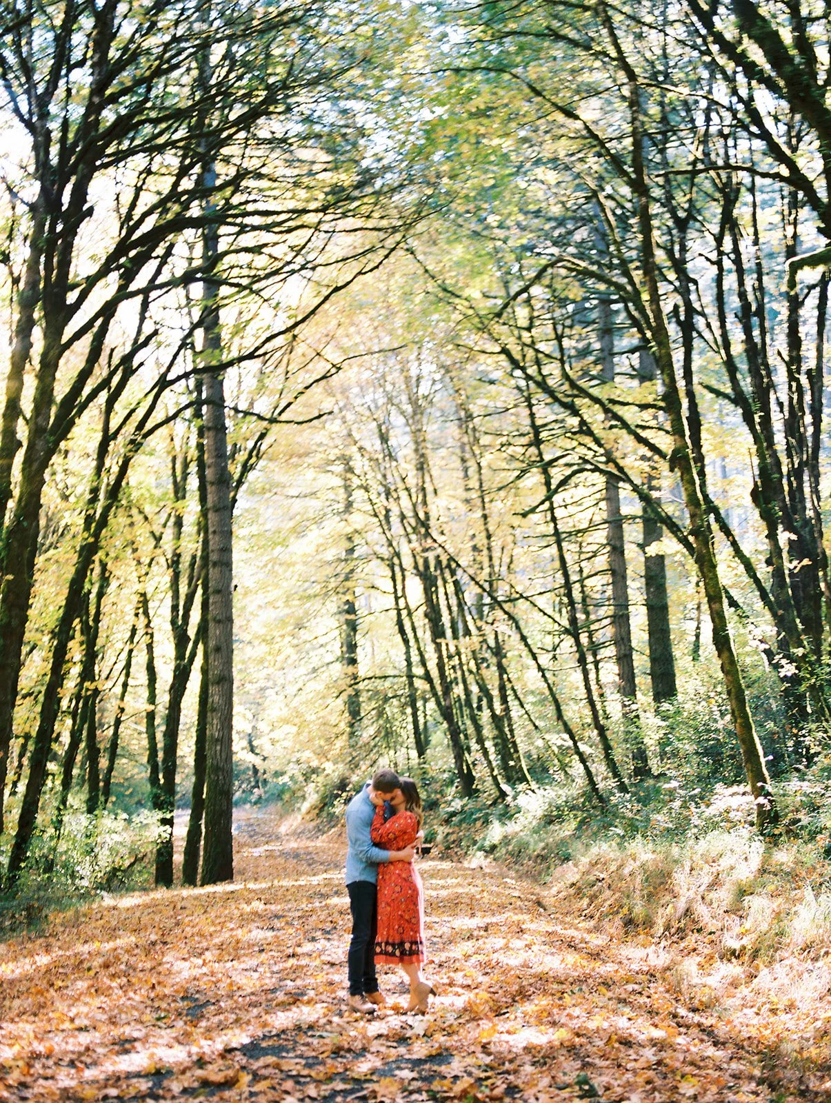 Couple embracing on a sunlit forest trail covered in autumn leaves during fall engagement photos Oregon