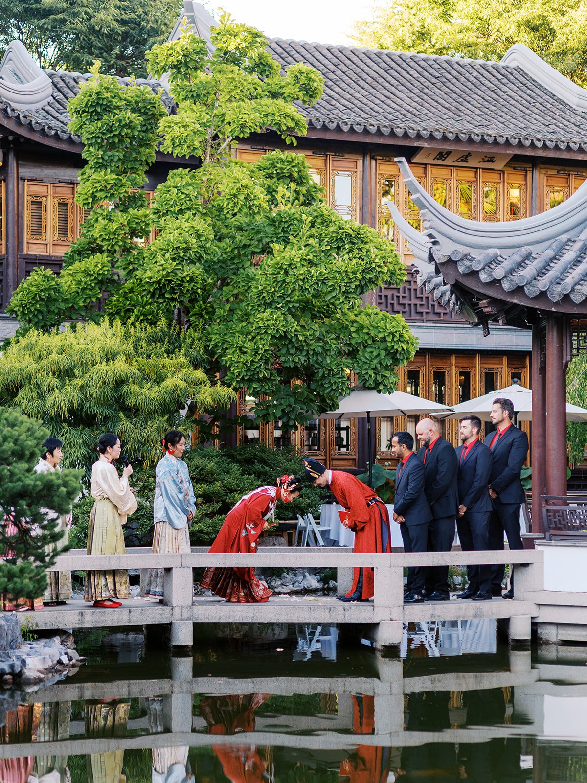The couple in red robes bow deeply together on the stone bridge during a traditional ceremony ritual, attendants in hanfu and groomsmen in black suits looking on, the garden reflected in the lake.