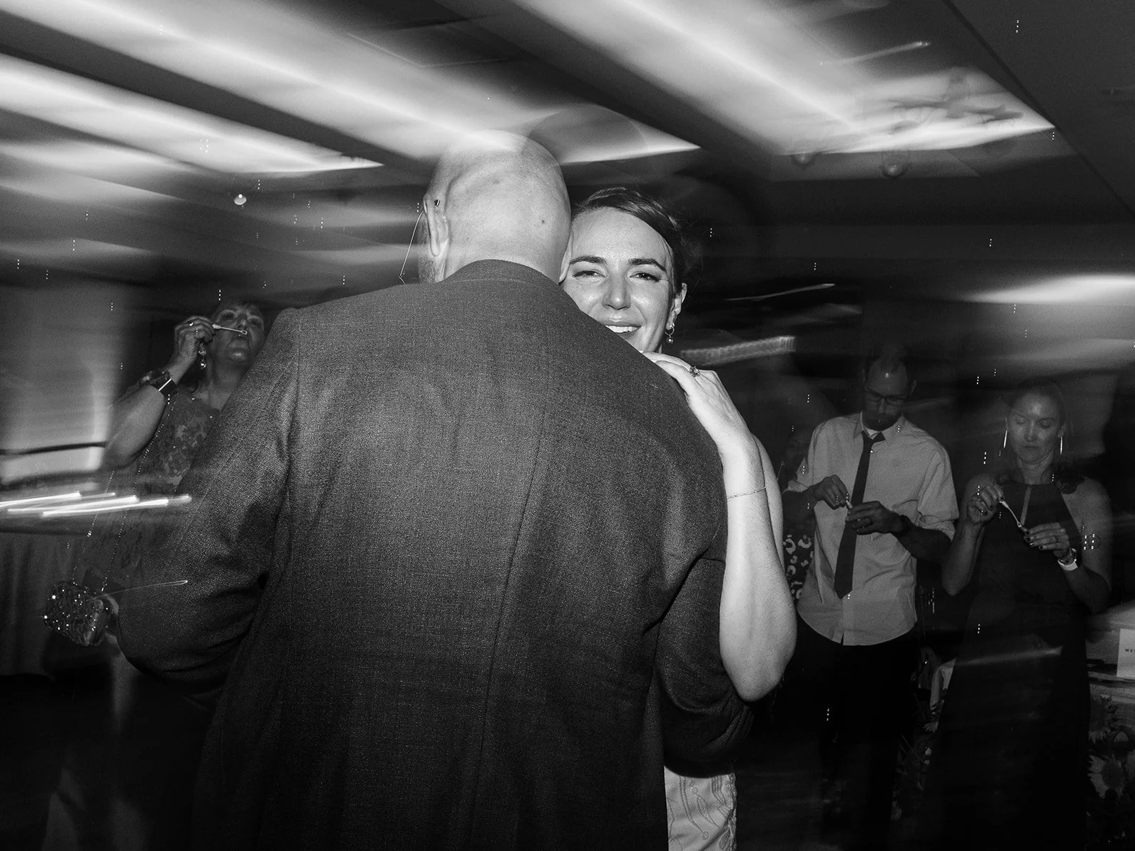 Black and white close-up of the bride smiling warmly over the groom's shoulder as they slow dance, guests blowing bubbles behind them