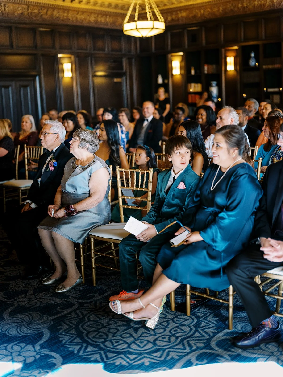 Wedding guests seated in gold chairs watching ceremony in an elegant indoor venue.