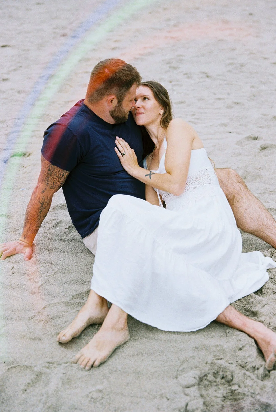 Couple seated on the beach, woman resting against man while looking up at him.