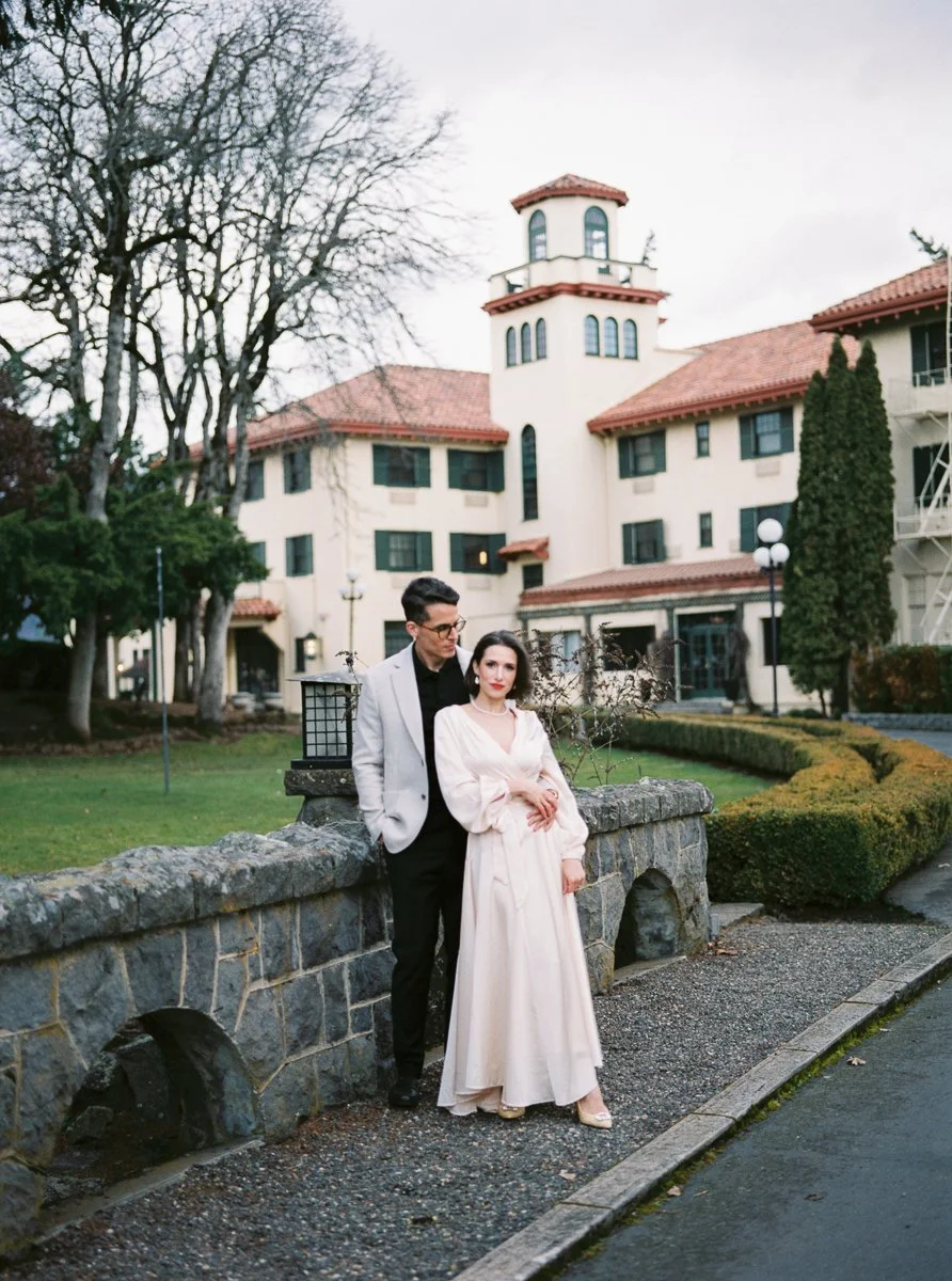 Couple posing by stone wall outside Spanish-style building, woman in cream dress, man in light blazer and black pants