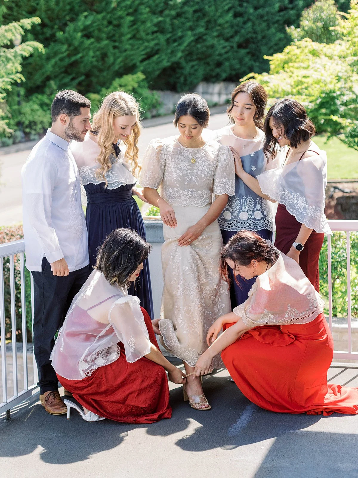 Bridesmaids in traditional Filipino attire helping the bride fasten her heeled sandals outdoors
