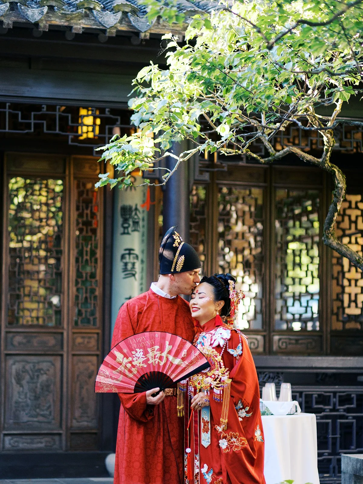 Groom pressing a kiss to the bride's forehead in front of an illuminated carved pavilion doorway at Lan Su Chinese Garden