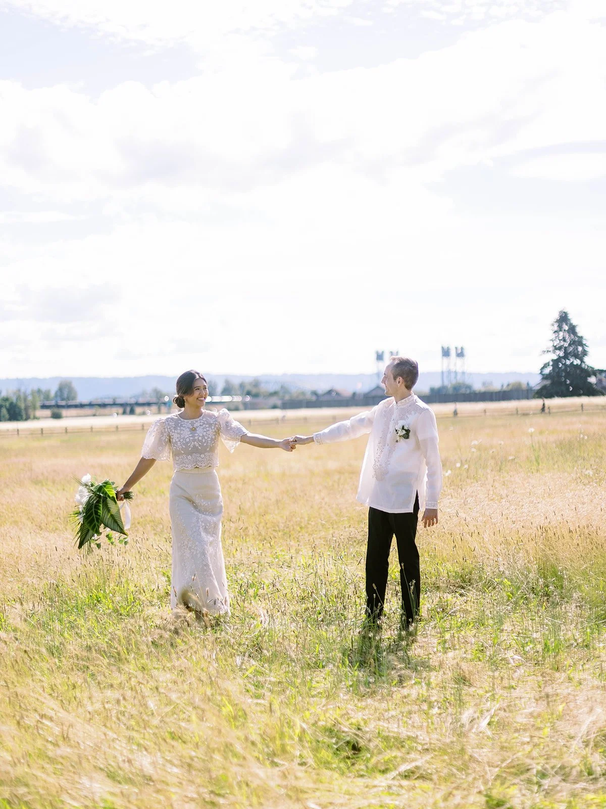 Bride and groom holding hands at arm's length in a sunlit golden field, smiling at each other with Pearson Airfield in the background