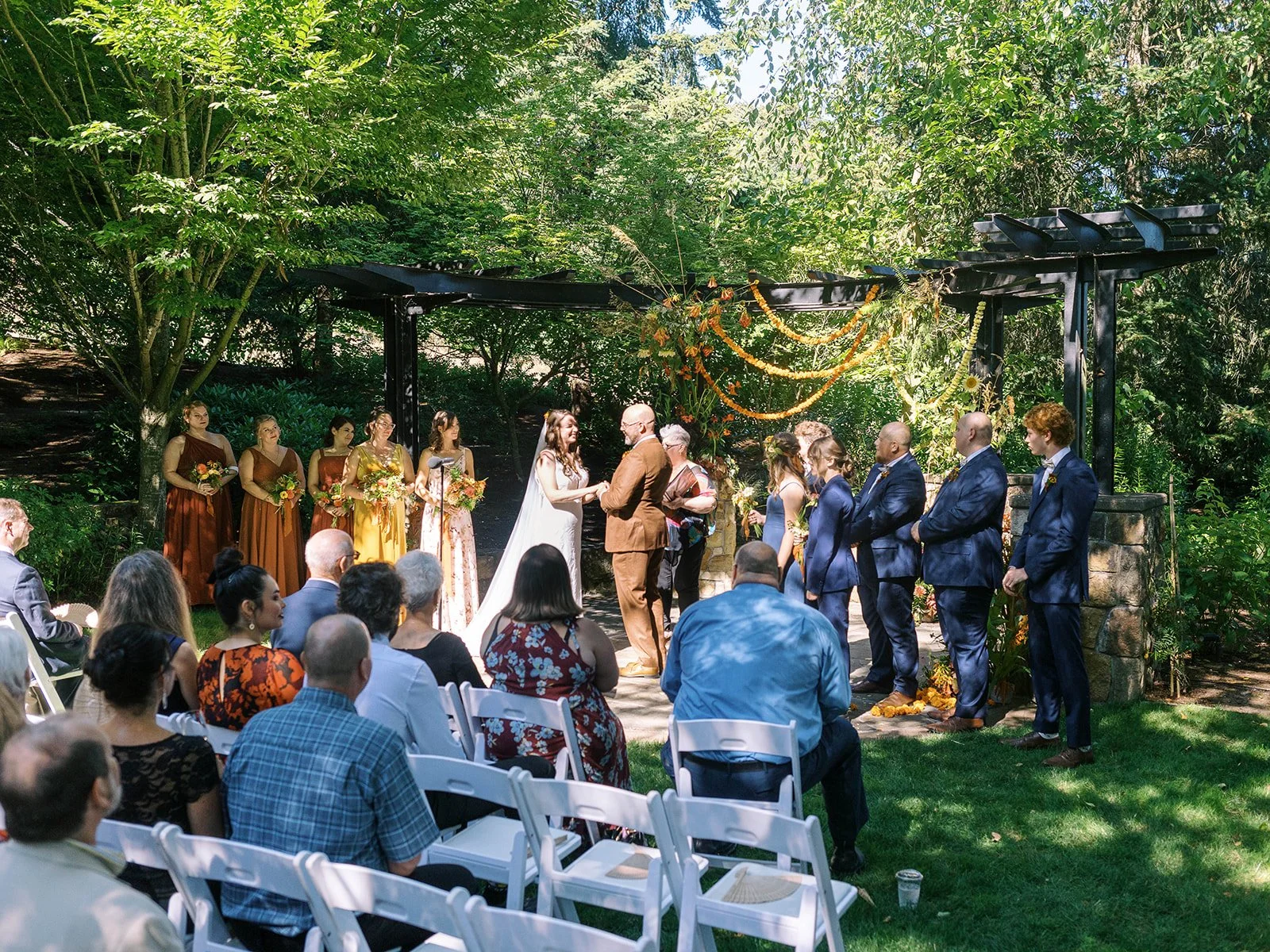 Bride and groom hold hands during ceremony under a shaded arbor with guests seated on both sides.