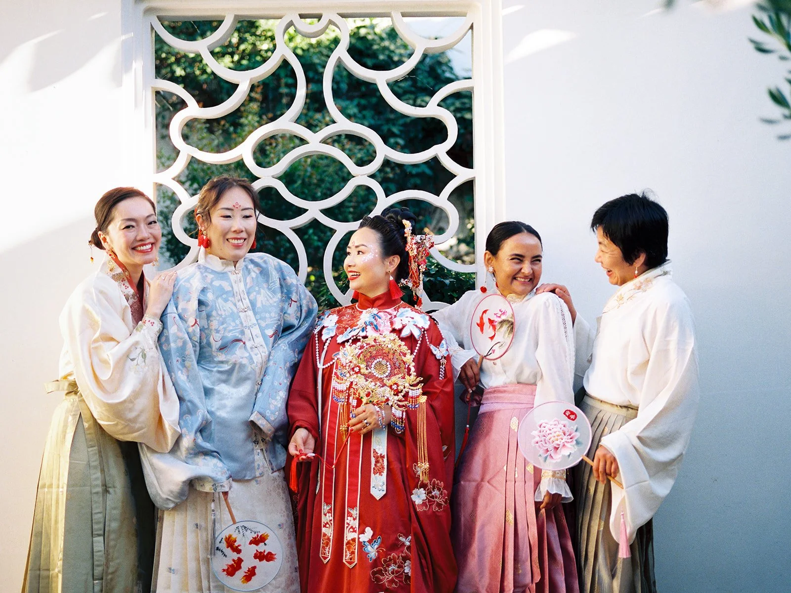 Bride in red qun kwa surrounded by four attendants in hanfu, all laughing together in front of a decorative lattice window at Lan Su Chinese Garden