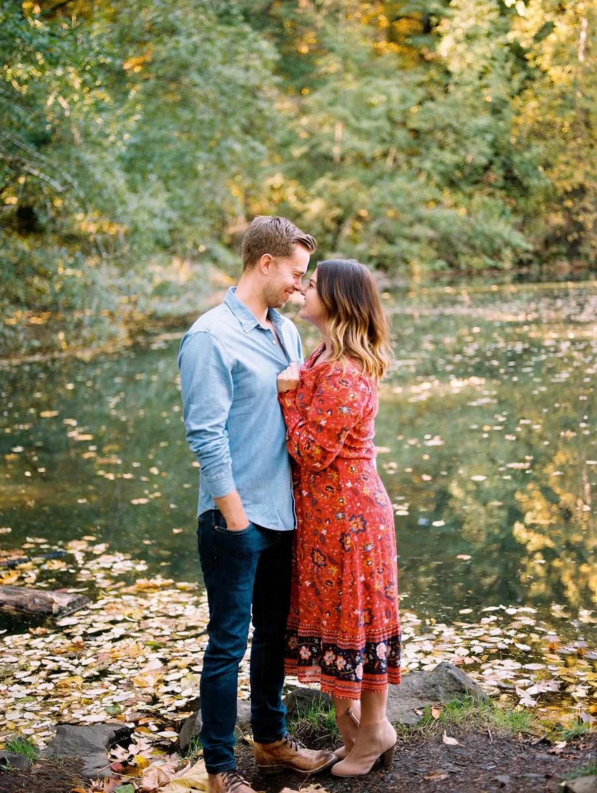Couple standing close by a calm river surrounded by trees with fall leaves during fall engagement photos Oregon