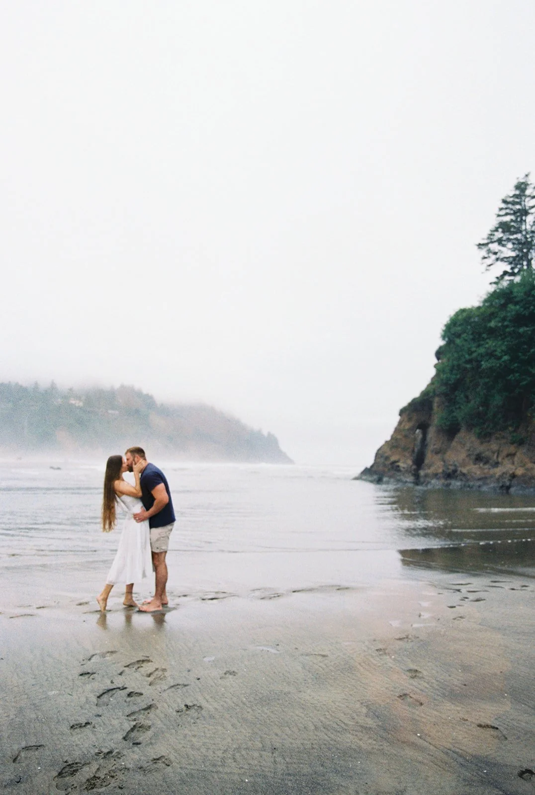 Couple kissing on wide, empty beach with soft fog, footprints in sand, and distant cliffs.