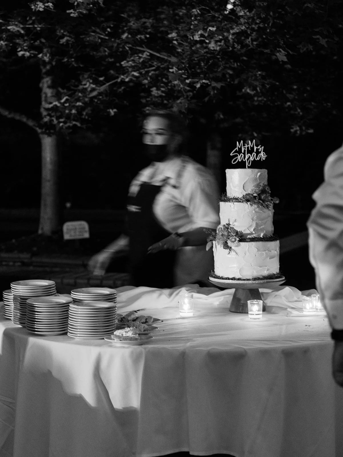 Wedding cake displayed on table with candles and stacked plates during evening reception