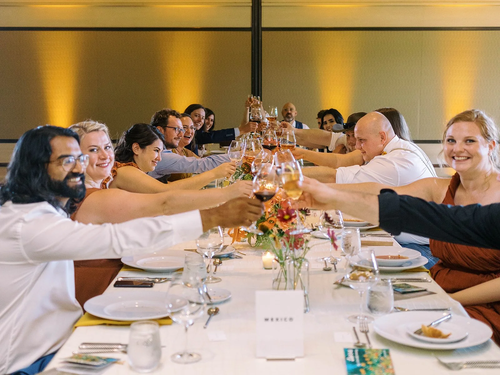 Wedding guests at a long reception table raising wine glasses in a toast, laughing and smiling, with a table card reading "Mexico" visible in the foreground