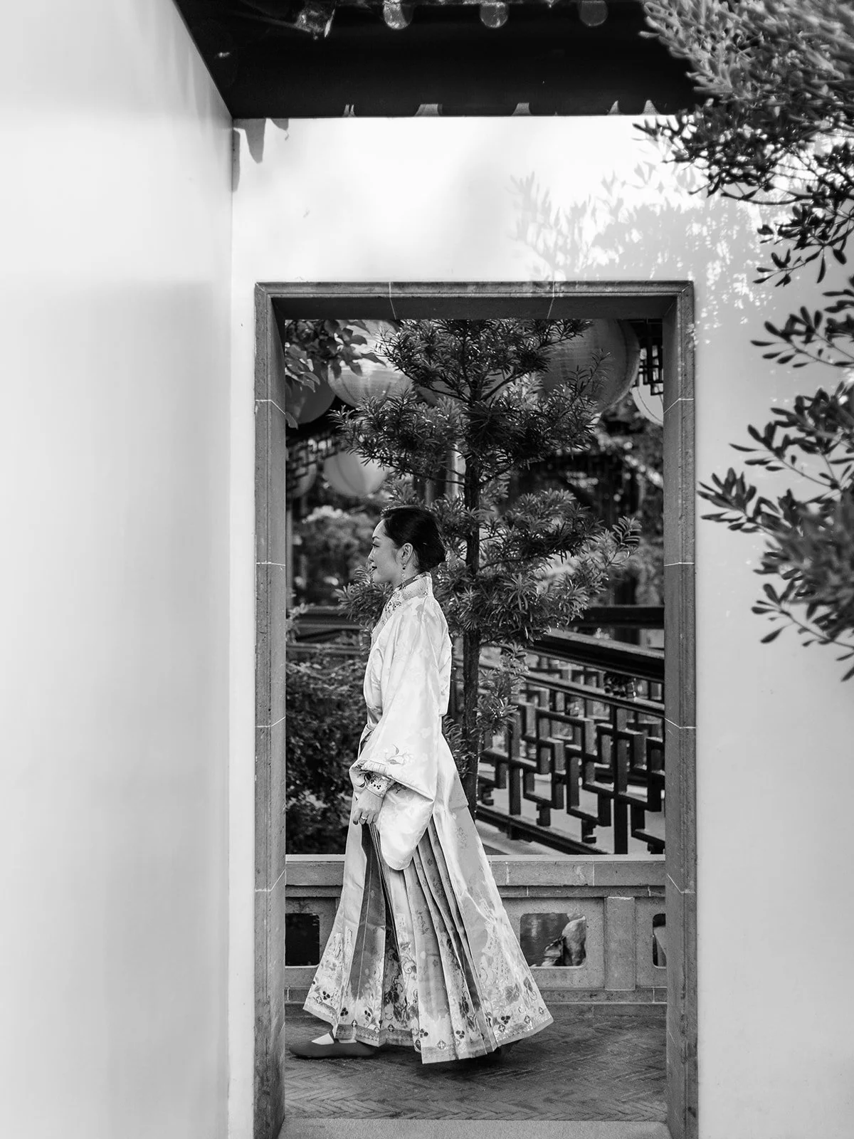 Black and white image of the bride in flowing white hanfu seen in profile through a rectangular moon gate doorway, with garden lattice and pine tree beyond.