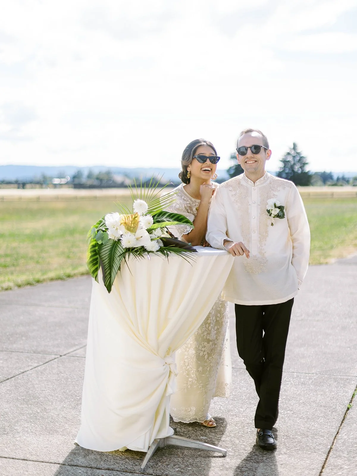 Bride laughing while leaning on a tropical floral-decorated cocktail table beside the groom on the airfield tarmac
