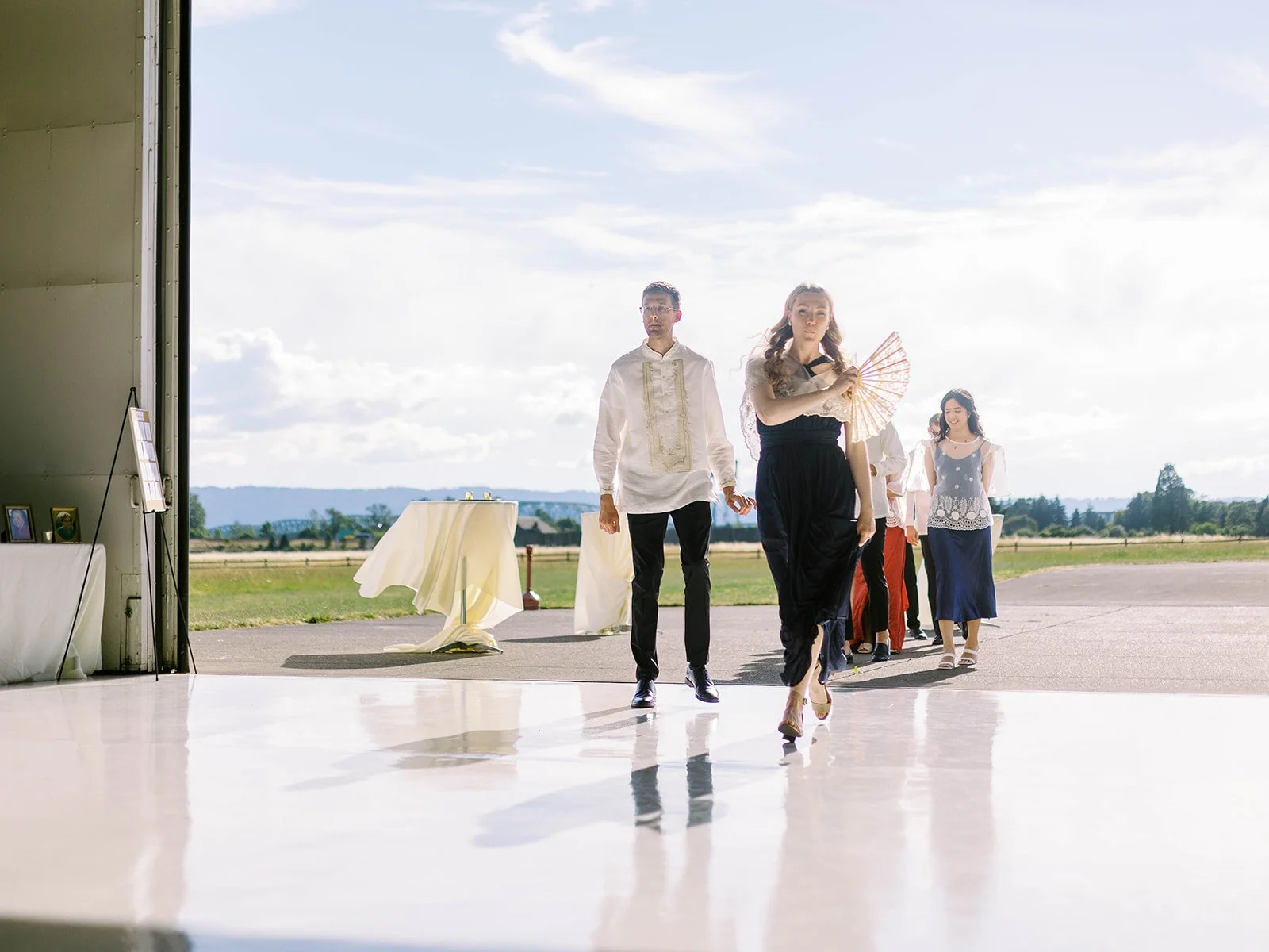 Wedding party members processing in a line through the bright hangar doors onto the reflective hangar floor
