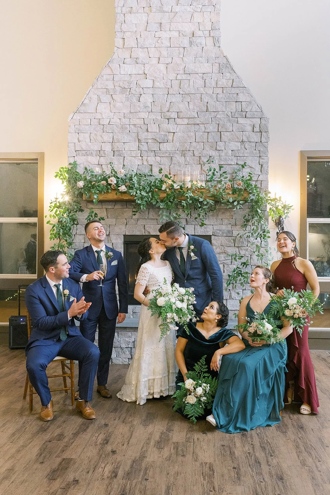 Bride and groom kiss as their wedding party gathers around them in front of the floral stone fireplace.