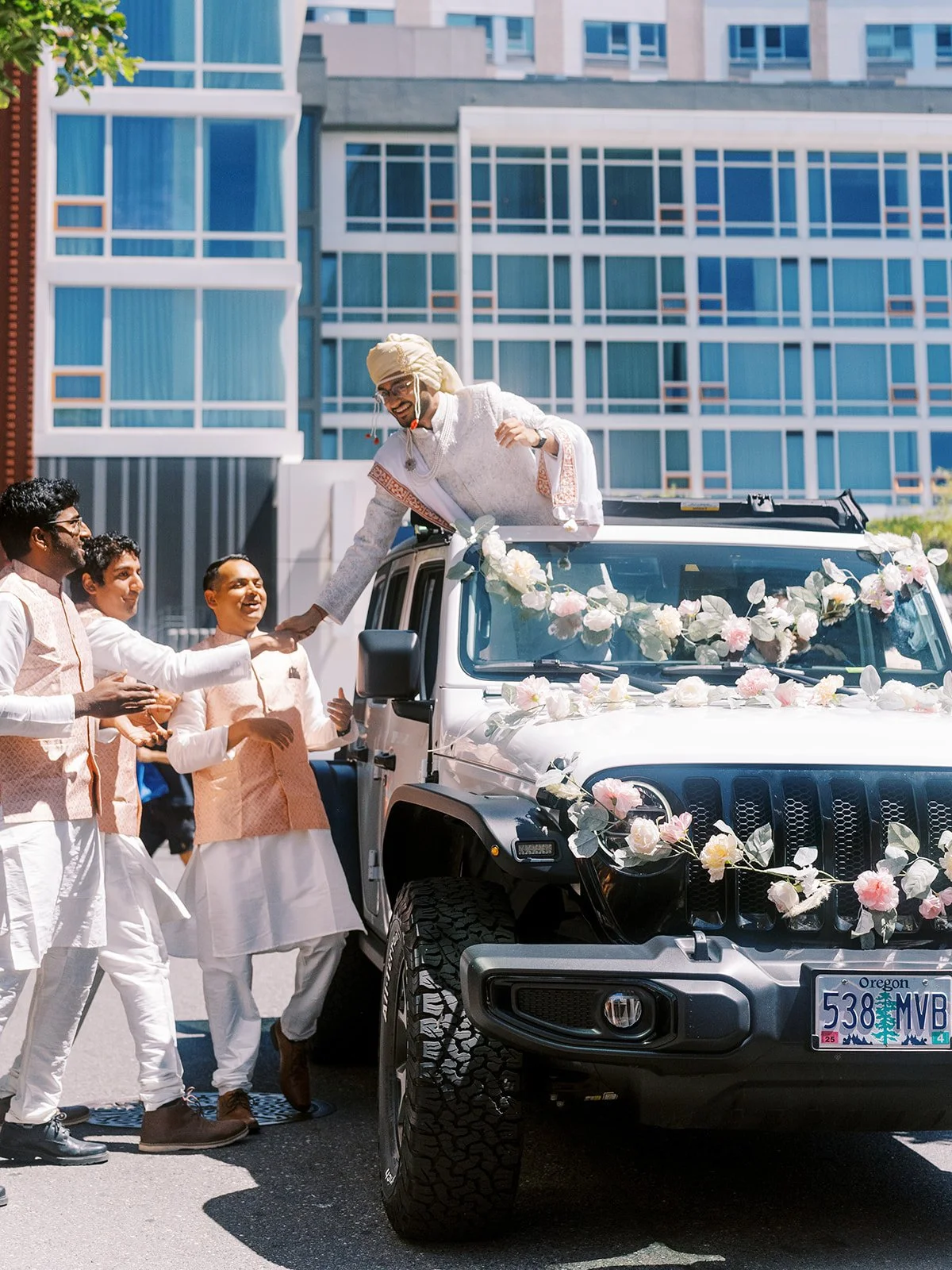 Groom in white sherwani and turban greeting friends from flower-decorated wedding car during baraat.