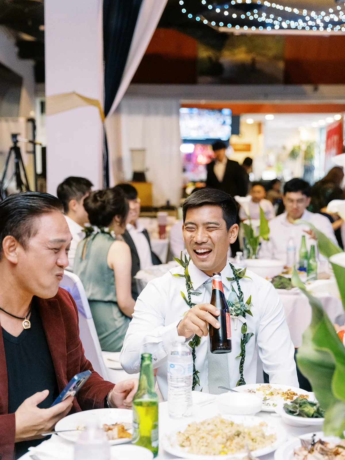 Two men laughing together at a wedding reception table, one wearing a green maile lei and holding a beer bottle, string lights glowing overhead