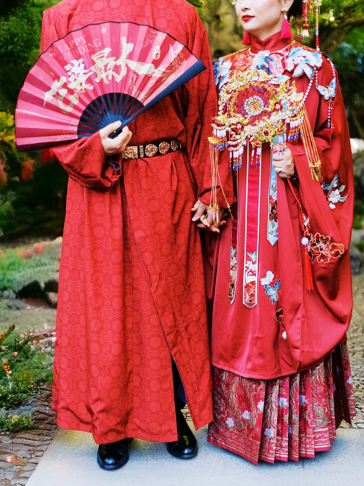 Detail shot of the bride's embroidered qun kwa and groom's red robe from the waist down, hands clasped together between them with the gold bridal bouquet visible