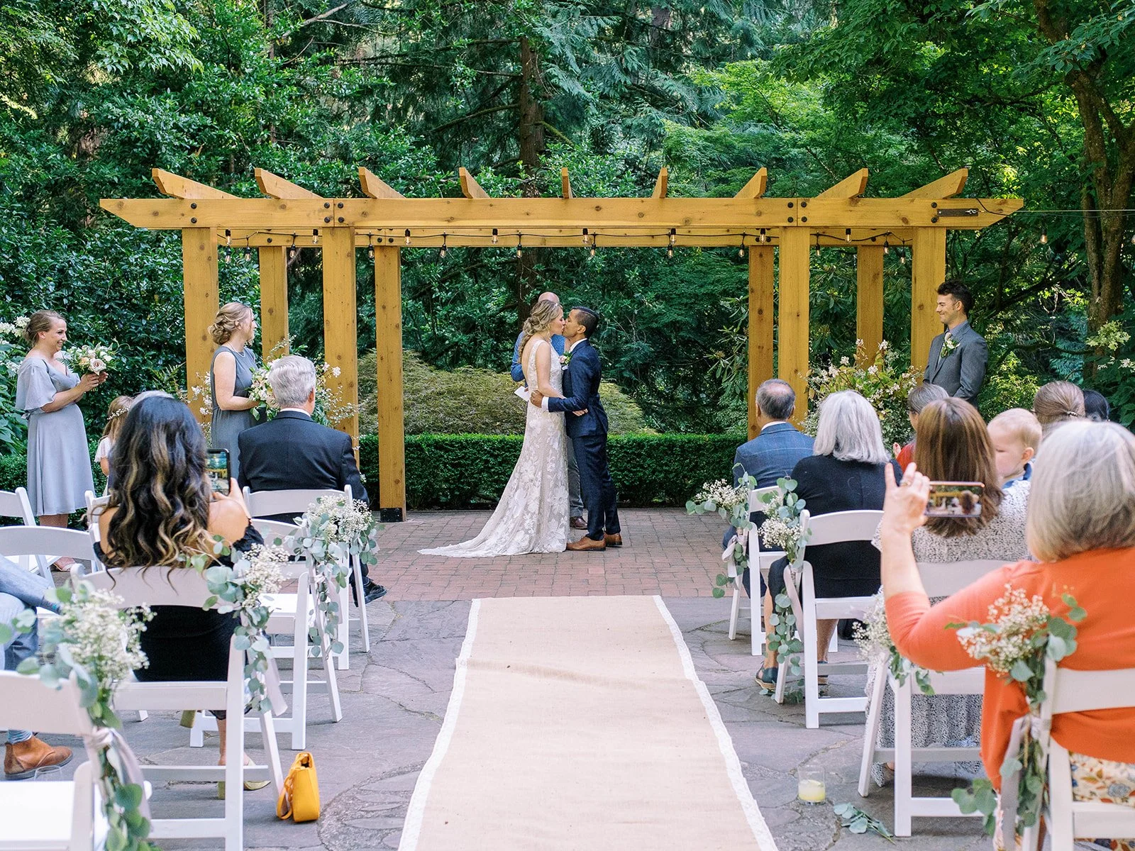 Bride and groom kiss beneath a wooden pergola as wedding guests watch from the aisle.