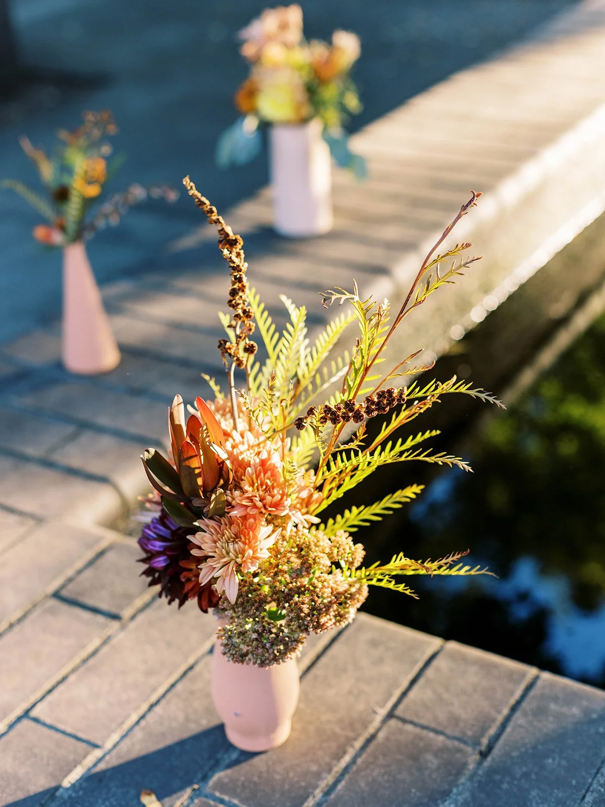 Small peach vases filled with warm-toned flowers arranged along the edge of a reflecting pool.