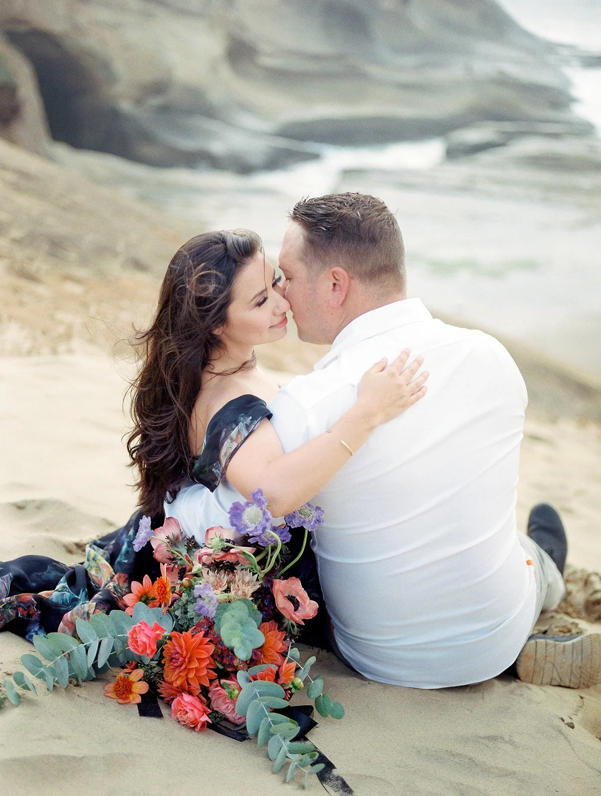 Couple sitting on sandy beach cliff, embracing with ocean and rock formations in soft coastal light