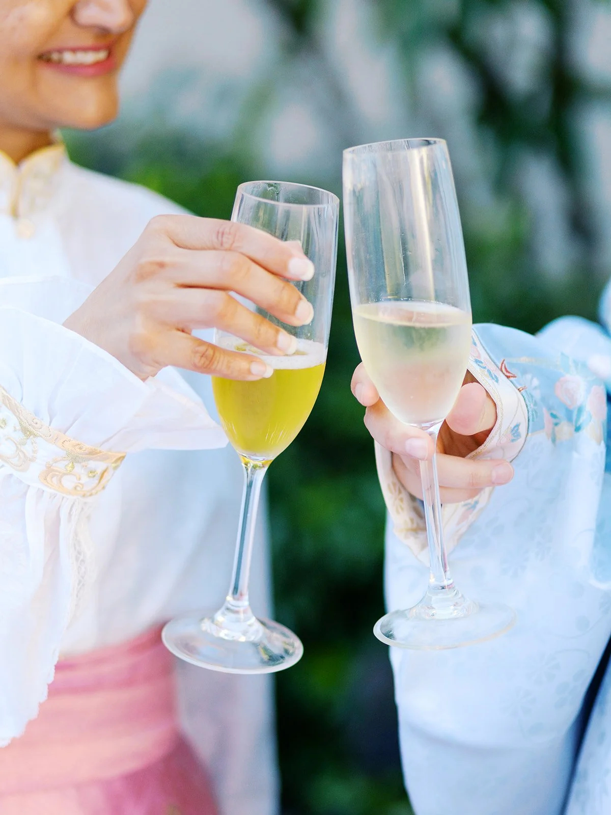 Close-up of two hands in traditional Chinese garments clinking champagne flutes against a soft green bokeh background.