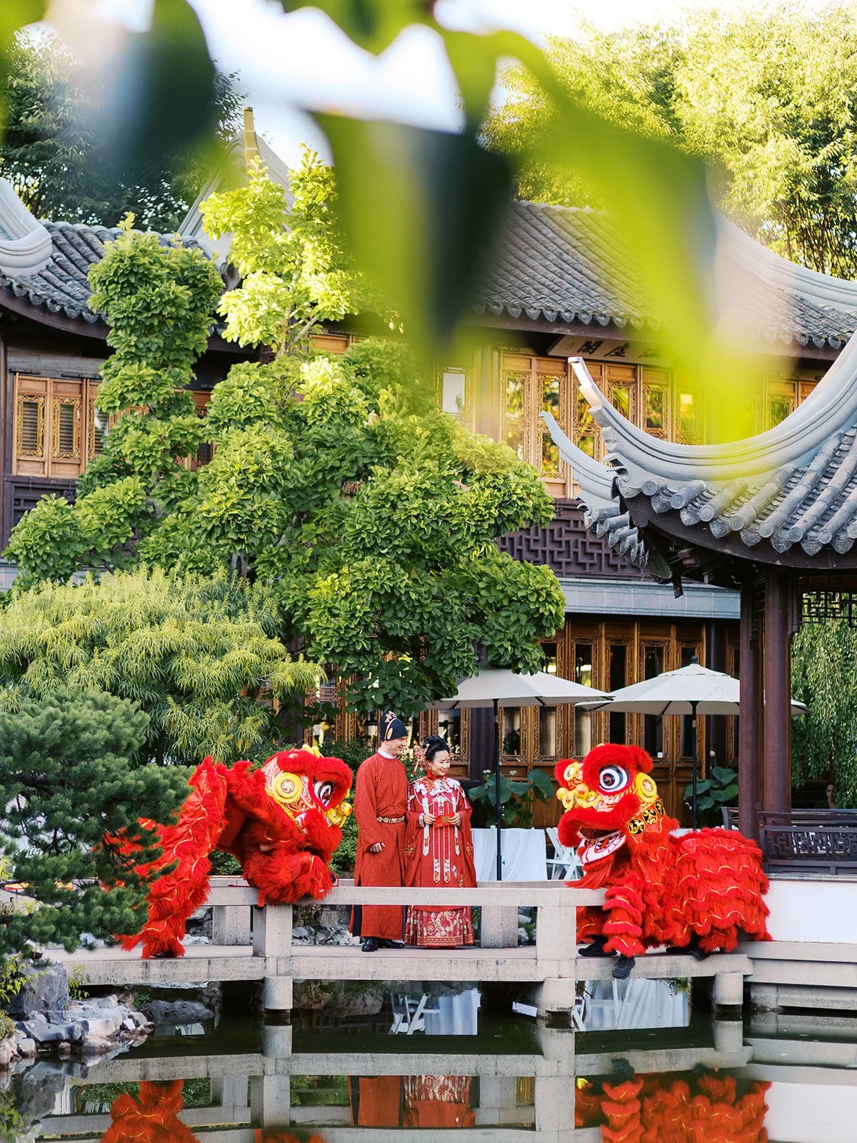 The couple in red wedding robes stand together on the bridge, two red lion dance costumes bowing toward them on either side, the lush garden and curved pavilion roofline behind.