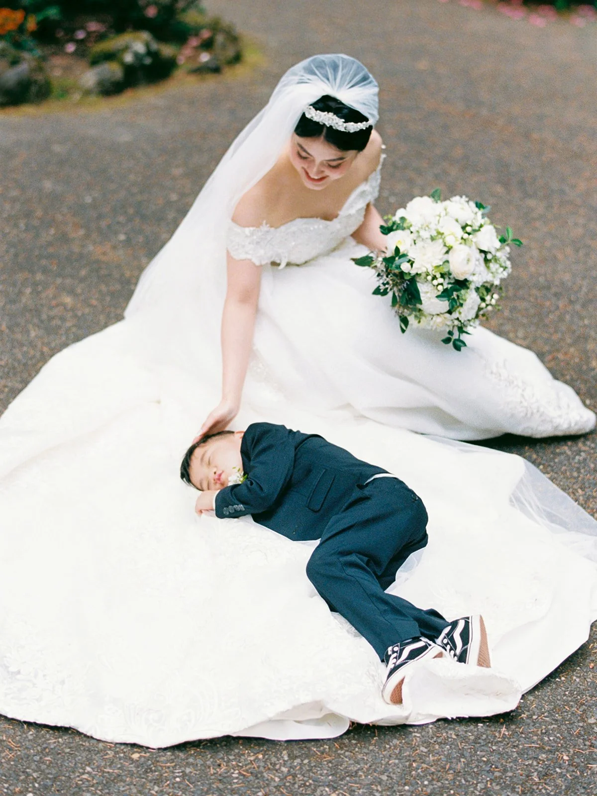 Bride in a lace ball gown and veil kneeling on the ground at The Grotto, smiling down at the ring bearer who has fallen asleep across the skirt of her gown